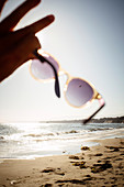 Close up of person on sandy beach, holding sunglasses towards the sun.