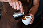 Close up of barista wearing brown apron pouring cappuccino.