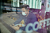 Mann mit Gesichtsmaske sitzt an einem Cafétisch und benutzt ein Mobiltelefon, Blick durch ein Fenster