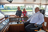 Paar plaudert mit Kapitän auf der Brücke von Flusskreuzfahrtschiff während einer Kreuzfahrt auf dem Rhein, nahe Andernach, Rheinland-Pfalz, Deutschland, Europa