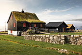 Schwarze Häuser mit roten Fenstern und Grasdach im Dorf Kirkjubøur auf Streymoy bei Sonne, Färöer Inseln\n