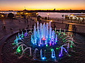 Luftaufnahme von beleuchtetem Musikbrunnen entlang Promenade am Ufer von Fluss Wolga in der Abenddämmerung, Astrachan, Bezirk Astrachan, Russland, Europa