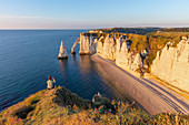 Eine Frau sitzt n der Alabasterküste und betrachtet den Felsbogen Porte d'Aval (Elefantenrüssel) und die Felsnadel Aiuille bei Étretat, Normandie, Frankreich