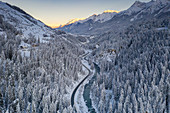 Sonnenaufgang über kurvenreiche Bergstraße und gefrorener Fluss im mit Schnee bedeckten Wald, Zernez, Kanton Graubunden, Schweiz, Europa