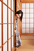 Glückliche verspielte junge Frau im Kimono, die hinter der Shoji-Tür späht
