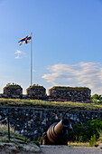Kanone mit Flagge, Naherholungsgebiet und Festungsanlage auf der Insel Suomenlinna vor Helsinki, Finnland