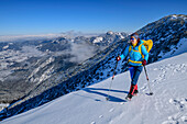 Woman hiking with snowshoes on her backpack goes over snow-covered mountain slope, Bavarian Alps in the background, Jägerkamp, Spitzing area, Bavarian Alps, Upper Bavaria, Bavaria, Germany