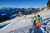 Man and woman hiking up over snow slope to Hochgern, Hochgernhaus in the background, Hochgern, Chiemgau Alps, Upper Bavaria, Bavaria, Germany