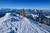 Man and woman hiking up over snow-covered slope to Hochgern, Kaiser Mountains and Chiemgau Alps in the background, Hochgern, Chiemgau Alps, Upper Bavaria, Bavaria, Germany
