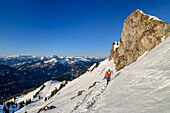 Mann und Frau beim Wandern steigen über Schneehang zur Rotwand auf, Felstürme im Hintergrund, Rotwand, Spitzinggebiet, Bayerische Alpen, Oberbayern, Bayern, Deutschland