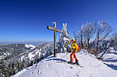 Woman on ski tour stands at the summit cross of the Predigtstuhl, Predigtstuhl, Chiemgau Alps, Upper Bavaria, Bavaria, Germany