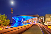 Blau beleuchtetes Gebäude der BMW Welt mit Olympiaturm im Hintergrund, München, Oberbayern, Bayern, Deutschland