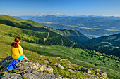 Woman while hiking sits on rock and looks down into the valley, Kamplnock, Nockberge, Nockberge-Trail, UNESCO Nockberge Biosphere Park, Gurktal Alps, Carinthia, Austria