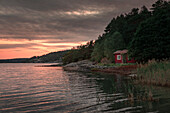 Rote Hütte am See mit Schilf auf Schäreninsel Orust an der Westküste von Schweden im Sonnenuntergang\n