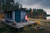 Frau sitzt vor Sauna am See in Lappland, Schweden\n