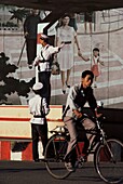 Mann, der mit seinem Fahrrad an einem chinesischen Soldaten vorbeifährt, der seinen Arm ausstreckt, während er vor einem Wandbild steht, das eine Mutter und eine Tochter zeigt, die sich an den Händen halten, China