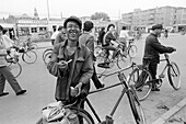 China, Datong, Man with crooked teeth smiling while pulling out cigarette and standing next to his bike, crowd walking and riding behind him