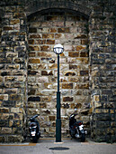 Two black motorbikes parked by a street lamp in front of a stone brick wall in the city