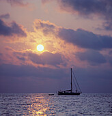 Silhouett einer Yacht auf ruhiger See und Sonne, die bei Sonnenuntergang zwischen Wolken scheint