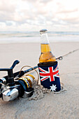 Bier im Stubbie-Halter mit australischer Flagge neben Angelrute im Sand am Strand