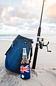 Beer in stubbie holder with Australian Flag, cooler bag and fishing rod on the beach on Australia Day