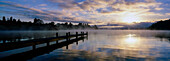 Panorama of mist raising from Lake Rotoiti at sunrise and wooden jetty stretching out from shore