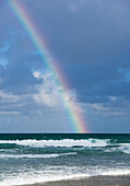 Regenbogen, der im stürmischen Himmel scheint, während Wellen auf Ufer rollen