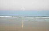 Der Mond steigt am Abendhimmel über dem Ozean auf und spiegelt sich im Sand