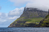 A few small houses on a cliff in front of a mountain backdrop. Gasadalur, Vagar, Faroe Islands.