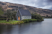 Kleine steinerne Kapelle mit Holztür am Seeufer. Berg im Hintergrund. Derreennacusha, Bealanageary, County Cork, Irland.