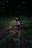 Woman standing on wooden fence in a forest holding a lamp in the dusk.