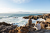 Frau und Teenager-Mädchen sitzen am felsigen Ufer und blicken aufs Meer.