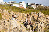 Mother and teenage daughter sitting on the rocks below luxury houses on a headland at De Kelders, South Africa
