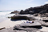 Teenager-Mädchen liegt auf dem Rücken auf Felsen über einem Sandstrand, Walker Bay Naturreservat, Südafrika