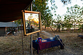 Ein Safaricamp, Zelte und Getränketisch, ein großer Spiegel an einer Zeltstange, drei Personen bei Sonnenuntergang, Okavango-Delta, Botswama, Südafrika