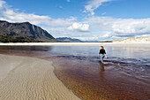 A teenage girl wading through a water channel on a wide sandy beach, Grotto Beach, South Africa