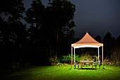 A small gazebo in a garden, a table and seat and lights at night