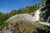 Frau beim Wandern steht auf Brücke und blickt auf Rutor-Wasserfälle mit Regenbogen, Rutorfälle, Rutorgruppe, Grajische Alpen, Aosta, Italien
