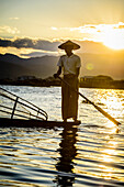 A fisherman standing on his boat fishing on Lake Inle at dusk.