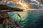 Ein junger Mann taucht bei Sonnenuntergang in das ruhige Meer an der nördlichen felsigen Küste von Kauai, Hawaii, Vereinigte Staaten von Amerika, Pazifik