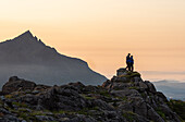 Ein Paar steht auf einem felsigen Gipfel und beobachtet einen Bergsonnenuntergang mit Sgurr nan Gillean im Hintergrund, Isle of Skye, Innere Hebriden, Schottland, Vereinigtes Königreich, Europa