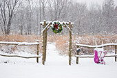 Rustikaler Zaun und Laube mit Weihnachtskranz und lila Schubkarre in der Nähe der Prärie im Winter, Marion County, Illinois