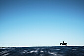 Mädchen reitet Paint Horse in der Ferne auf einem verschneiten Bergrücken unter blauem Himmel