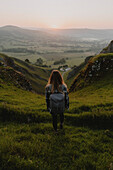 Female hiker on grassy hill watching sunrise in distance, Winnats Pass, Castleton, England