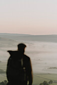 Blurred male hiker looking at scenic, foggy sunrise landscape, Winnats Pass, Castleton, England
