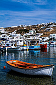 Mykonos, Griechenland. Orange-weißes Boot schwimmt im Wasser mit anderen bunten Booten und weiß getünchten Gebäuden auf dem Hügel