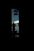 Eleganter Kellner in Silhouette, der mit seinen Händen in seinen Hüften auf einer Terrasse mit Sonnenschirm und einer Lampe in der Schweiz steht.