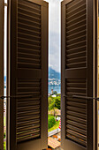 Fensteransicht mit Fensterladen mit Turm im Stadtbild und Berg und Luganersee an einem bewölkten Tag im Tessin, Schweiz.