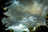 Gewitterwolken nähern sich, während ein Regenbogen bei Sonnenuntergang von Sonnenstrahlen beleuchtet wird.