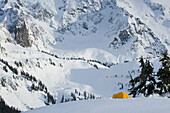 A small yellow tent pitched in deep snow on a slope, view of the steep slopes of the mountains.